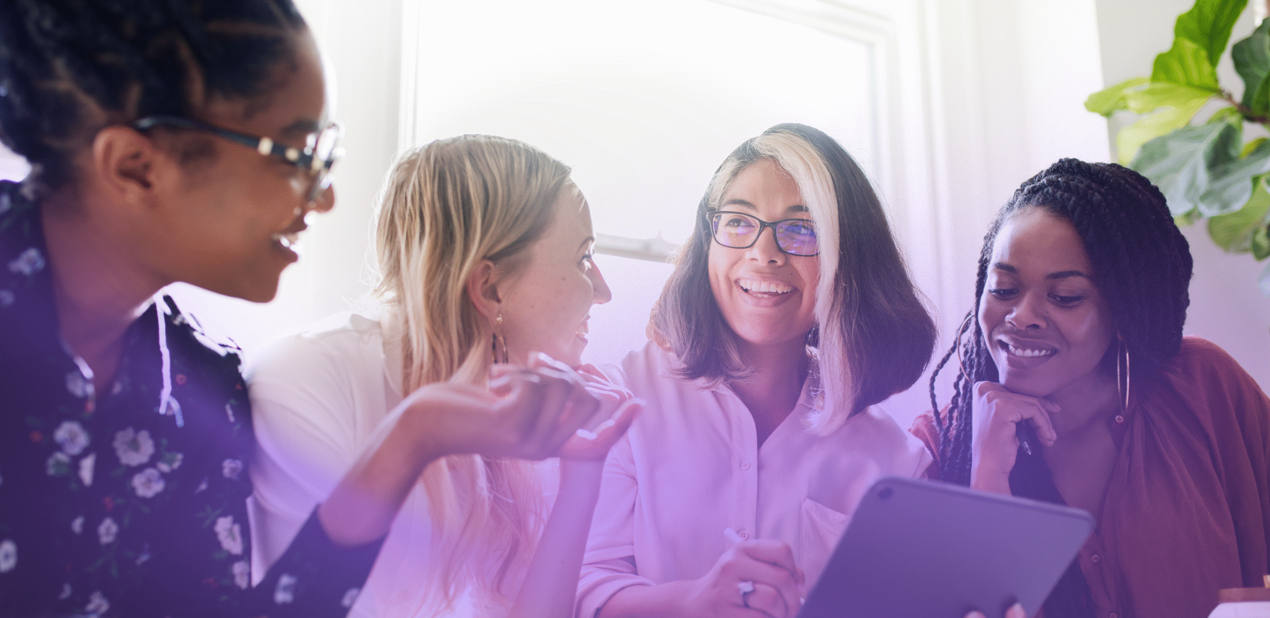 Four diverse women sitting together in a bright room, smiling and discussing something on a digital tablet.